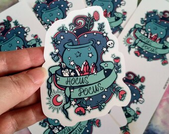 May include: A sticker featuring a teal cauldron with stars, crystals, and flowers, with the words "Hocus Pocus" on a banner. The design includes a crescent moon, skulls, and a butterfly. Several stickers are visible.
