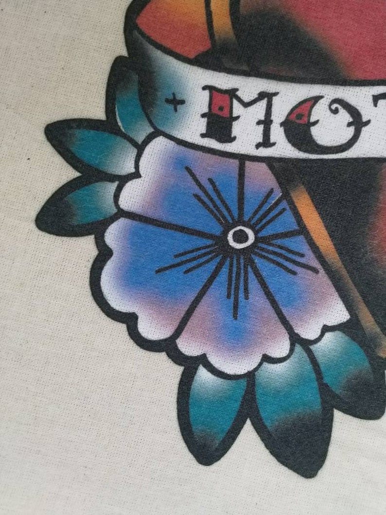 May include: A traditional tattoo design featuring a red heart with a banner that reads "MOM". The heart is surrounded by a blue flower with a white center and teal leaves.