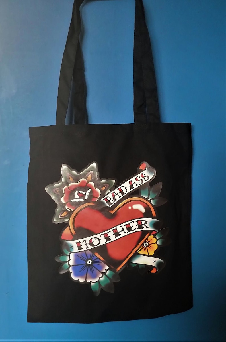 May include: Black tote bag with a heart-shaped tattoo design. The heart is red with a banner that reads "BAD ASS MOTHER". The design includes flowers and leaves. The bag has black handles.
