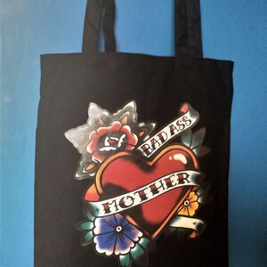 May include: Black tote bag featuring a heart-shaped tattoo design with the words "BAD ASS MOTHER" on a banner. The design includes roses, flowers, and leaves in red, blue, yellow, and green. The bag has two black handles.