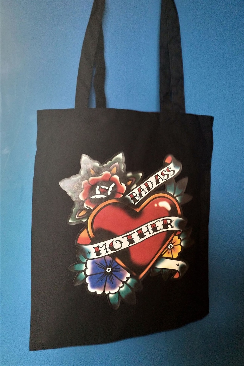 May include: Black tote bag featuring a heart-shaped tattoo design with the words "BAD ASS MOTHER" on a banner. The design includes roses, flowers, and leaves in red, blue, yellow, and green. The bag has two black handles.