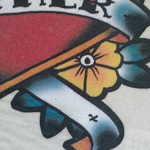 May include: A white t-shirt with a red heart and a banner that says "Together". The heart is outlined in black and has a yellow flower with a blue center. The banner is blue and white with a yellow outline.
