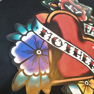 May include: Black tote bag featuring a colorful tattoo-style graphic. The design includes a red heart, flowers, and a banner with the words "MOTHER". The artwork has a vintage aesthetic with bold lines and vibrant colors.