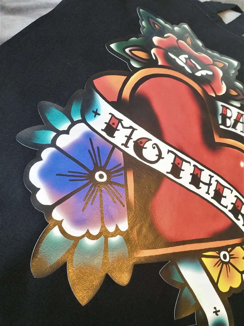 May include: Black tote bag featuring a colorful tattoo-style graphic. The design includes a red heart, flowers, and a banner with the words "MOTHER". The artwork has a vintage aesthetic with bold lines and vibrant colors.