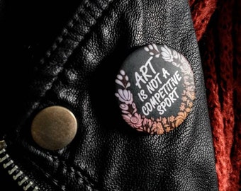 May include: A black button with a white floral border and the text "Art is not a competitive sport" in white lettering.