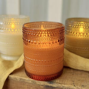 May include: Three cylindrical glass candle holders with a textured hobnail pattern. The center holder is amber, the left is clear, and the right is brown. Each contains a lit candle, casting a warm glow through the patterned glass surfaces.