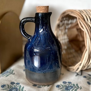 May include: A ceramic cruet with a glossy deep blue glaze on the upper portion and a matte charcoal gray base. The vessel features a curved handle, a narrow spout, and a light brown cylindrical cork stopper inserted into the top opening.