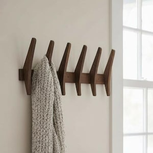 May include: A wooden coat rack with seven angled pegs is mounted on a beige wall. A light gray knitted scarf hangs from the rack. A window with white trim is in the background, and a distressed wooden bench sits below it.