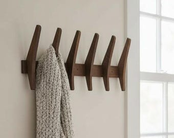 May include: A wooden coat rack with seven angled pegs is mounted on a beige wall. A light gray knitted scarf hangs from the rack. A window with white trim is in the background, and a distressed wooden bench sits below it.