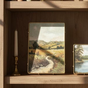 May include: A rectangular brass-framed print featuring a landscape painting of a winding dirt path through rolling green and golden hills under a soft sky. The frame is positioned on a wooden shelf next to a tall white taper candle in a brass holder.