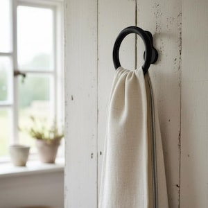 May include: A cream-colored kitchen towel with thin blue stripes hanging from a circular black metal ring mount. The towel is made of a woven fabric and features a simple hemmed edge. The ring is securely attached to a vertical white wooden surface.