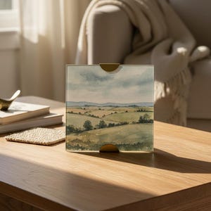 May include: A landscape watercolor print in a clear acrylic block with gold-colored accents. The artwork depicts rolling green hills under a cloudy sky. The block is displayed on a wooden coffee table, with a stack of books and a decorative bird.