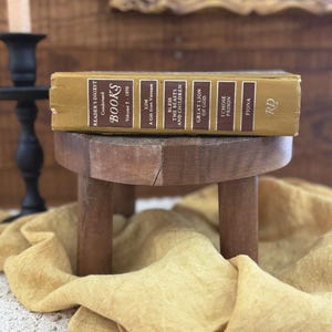 May include: A vintage Reader's Digest Condensed Books volume, featuring titles like "Kim" and "The Beasts and Children." The book, with a gold cover, rests on a small, rustic wooden stool. A yellow cloth and a candle holder are in the background.