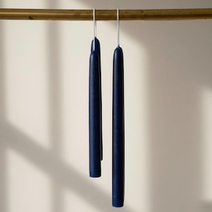 May include: Two long, slender taper candles in a deep navy blue color. The candles are suspended from a horizontal brass-colored rod by thin, silver-toned metal wires attached to the top of each candle, hanging vertically in a parallel arrangement.