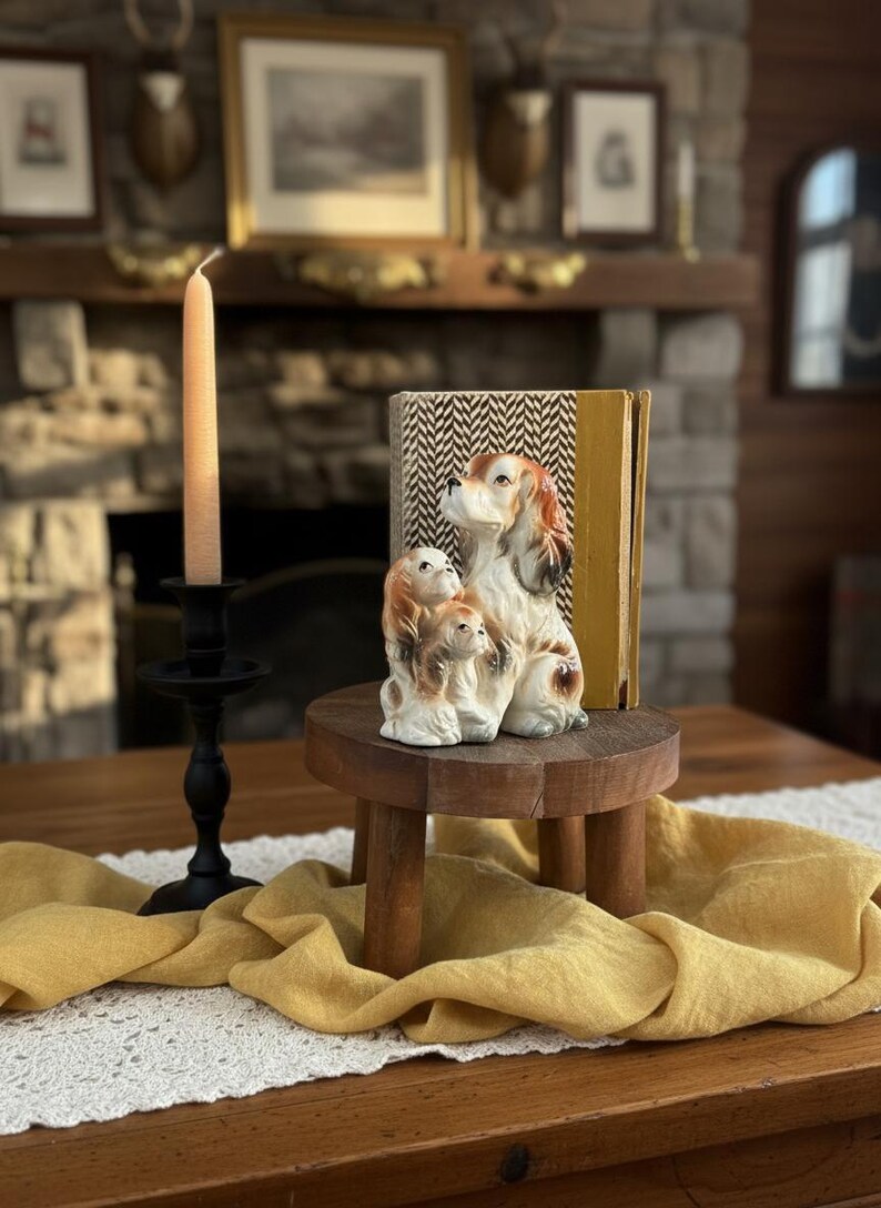 Puede incluir: Una figura decorativa de una familia de perros est&aacute; sobre un peque&ntilde;o pedestal de madera. La figura es marr&oacute;n, blanca y canela. Una vela alta de color melocot&oacute;n est&aacute; en un candelabro negro. Un camino de mesa amarillo con ribete de encaje.