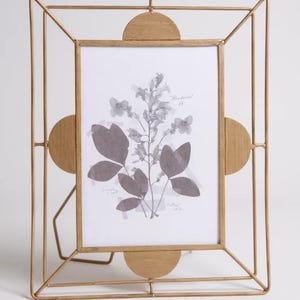 May include: A gold-colored metal picture frame with a geometric design. The frame holds a black and white print of botanical elements. The frame has a stand for tabletop display.