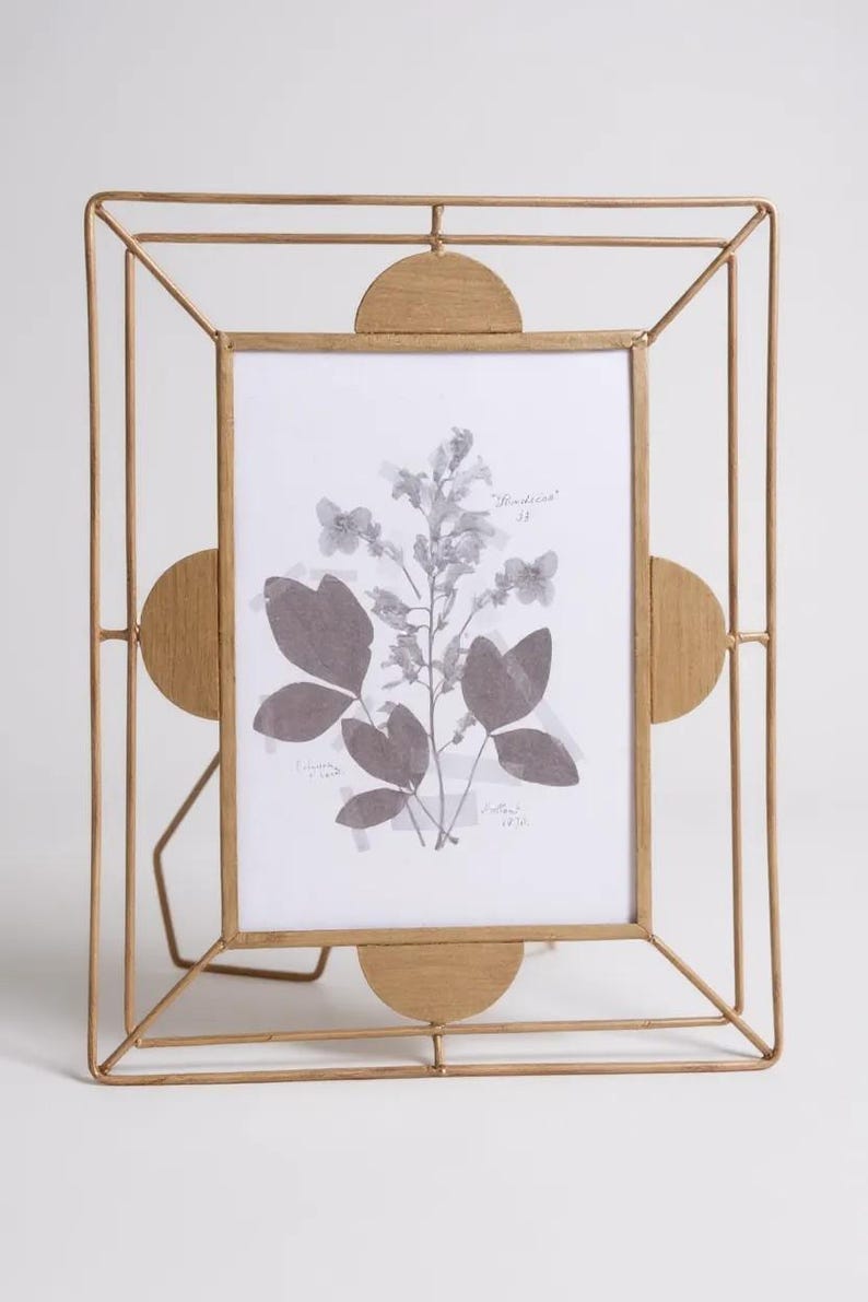 May include: A gold-colored metal picture frame with a geometric design. The frame holds a black and white print of botanical elements. The frame has a stand for tabletop display.