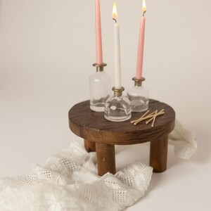 Puede incluir: Una peque&ntilde;a mesa redonda de madera sostiene tres velas encendidas en portavelas de cristal. Dos velas rosas y una blanca est&aacute;n encendidas. Un camino de mesa de encaje blanco y cerillas tambi&eacute;n est&aacute;n sobre la mesa.