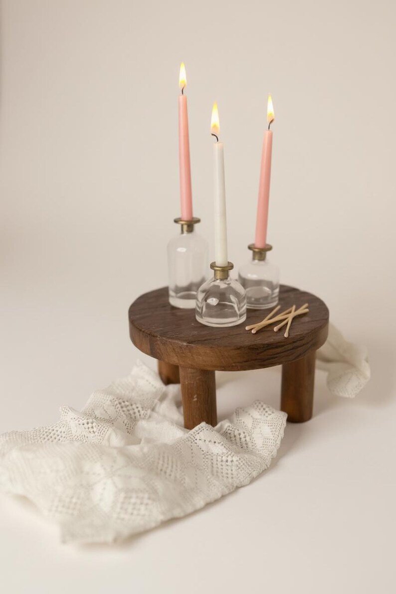 Puede incluir: Una peque&ntilde;a mesa redonda de madera sostiene tres velas encendidas en portavelas de cristal. Dos velas rosas y una blanca est&aacute;n encendidas. Un camino de mesa de encaje blanco y cerillas tambi&eacute;n est&aacute;n sobre la mesa.