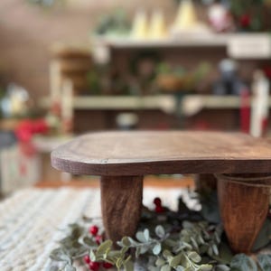 May include: A small, rectangular wooden riser with a smooth, dark brown finish and four sturdy, tapered legs. The riser is positioned over a decorative arrangement of green eucalyptus leaves and small red berries on a textured, woven fabric mat.