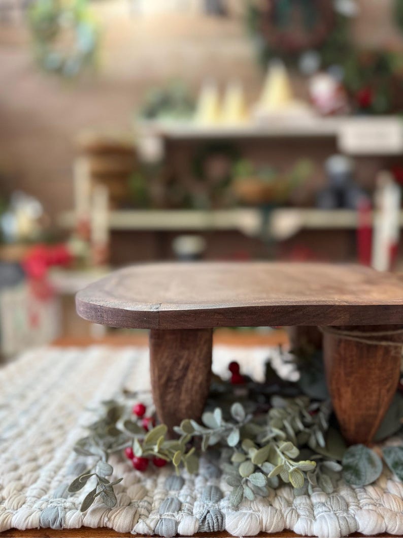 May include: A small, rectangular wooden riser with a smooth, dark brown finish and four sturdy, tapered legs. The riser is positioned over a decorative arrangement of green eucalyptus leaves and small red berries on a textured, woven fabric mat.