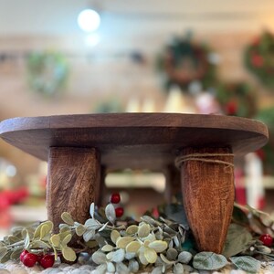 May include: A round, dark brown wooden riser stand featuring three thick, sturdy legs with a visible wood grain texture. The stand is positioned above a decorative arrangement of green foliage and small red berries on a textured white fabric base.