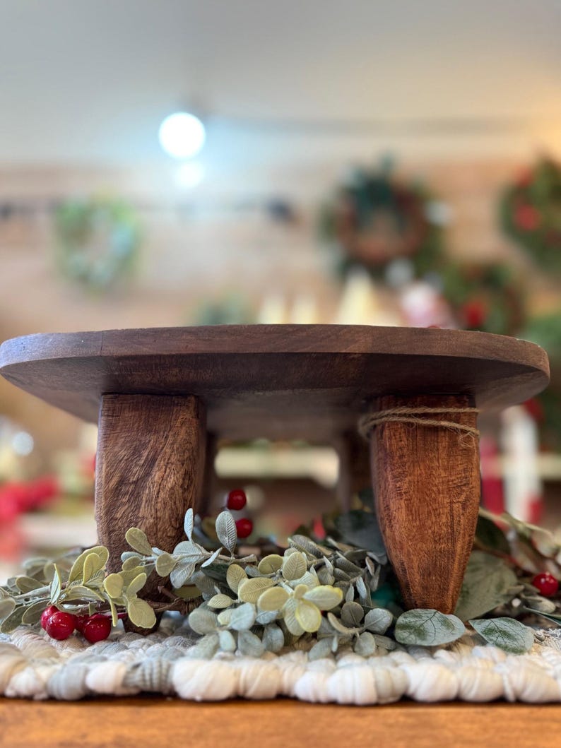 May include: A round, dark brown wooden riser stand featuring three thick, sturdy legs with a visible wood grain texture. The stand is positioned above a decorative arrangement of green foliage and small red berries on a textured white fabric base.