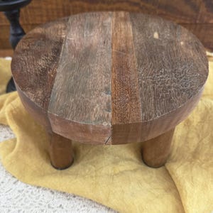 Puede incluir: Un peque&ntilde;o soporte redondo de madera para plantas con tres patas cortas. La mesa est&aacute; hecha de varias tablas de madera con diferentes tonos marrones y un acabado ligeramente envejecido. El soporte est&aacute; sobre una tela amarilla.