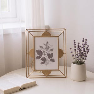 May include: A gold metal picture frame with a botanical print, a small white vase with purple flowers, and an open book on a white table. The frame has a geometric design and rounded accents.