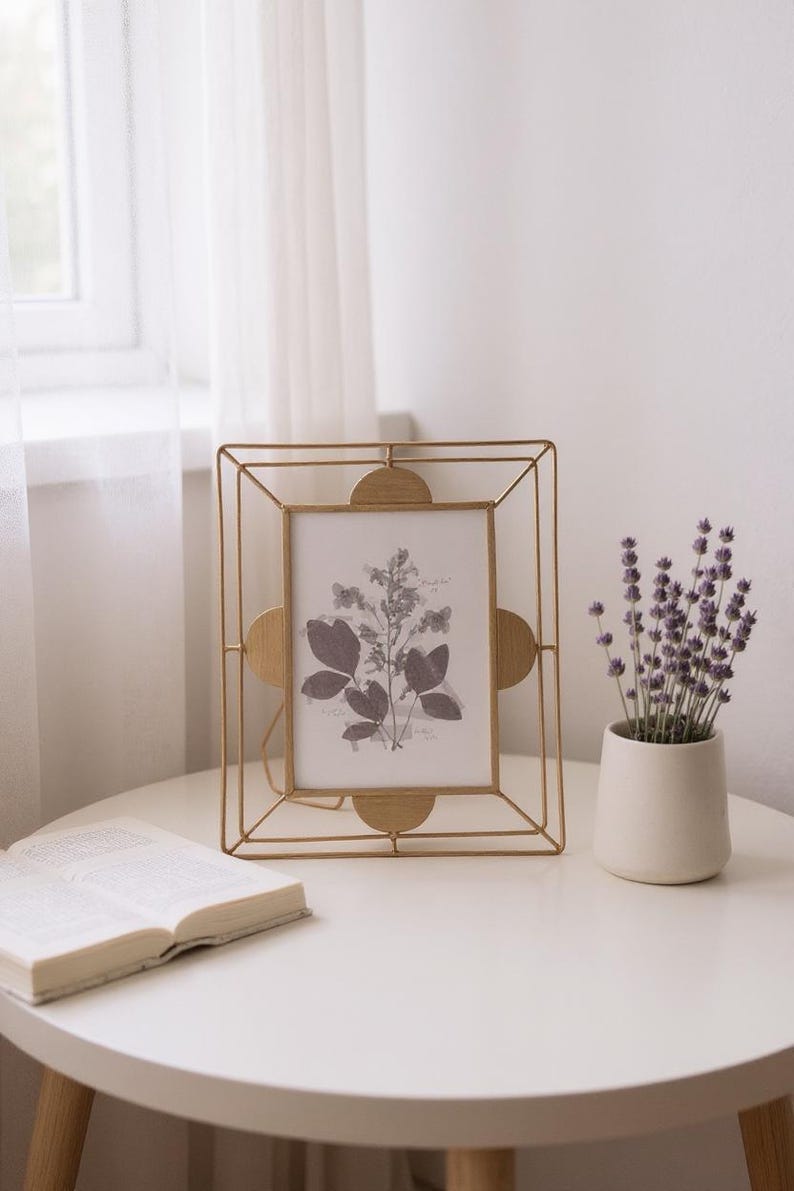 May include: A gold metal picture frame with a botanical print, a small white vase with purple flowers, and an open book on a white table. The frame has a geometric design and rounded accents.