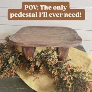 May include: A dark brown wooden pedestal with a square top featuring rounded corners and four short, thick legs. The text "POV: The only pedestal I'll ever need!" is displayed in white letters on a brown rectangular background above the object.