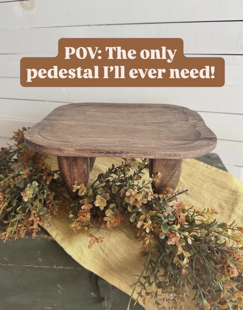 May include: A dark brown wooden pedestal with a square top featuring rounded corners and four short, thick legs. The text "POV: The only pedestal I'll ever need!" is displayed in white letters on a brown rectangular background above the object.