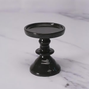 May include: A black ceramic candle holder with a round top and a pedestal base. The candle holder has a glossy finish and is designed to hold a single candle. The overall design is simple and elegant.