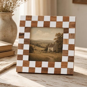 May include: A square picture frame featuring a checkerboard pattern of alternating natural brown wood and solid white resin tiles. The frame holds a landscape print depicting a stone house in a grassy field with rolling hills in the distance.