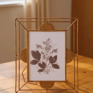 May include: A gold-colored metal picture frame with a rectangular opening, featuring a botanical print. The frame has a geometric design with rounded accents on the sides. The print showcases pressed flowers and leaves.