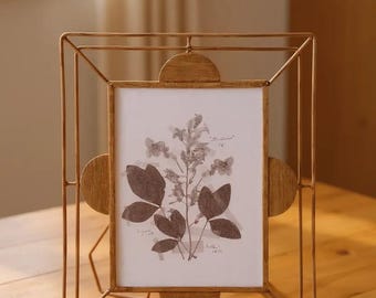 May include: A gold-colored metal picture frame with a rectangular opening, featuring a botanical print. The frame has a geometric design with rounded accents on the sides. The print showcases pressed flowers and leaves.