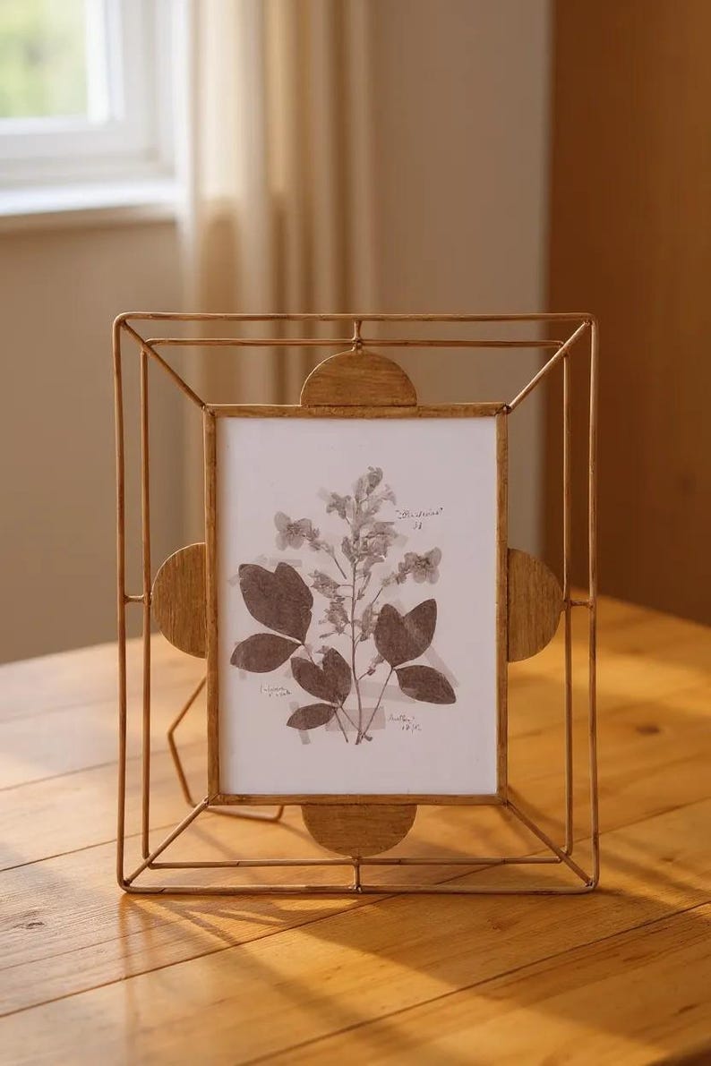 May include: A gold-colored metal picture frame with a rectangular opening, featuring a botanical print. The frame has a geometric design with rounded accents on the sides. The print showcases pressed flowers and leaves.