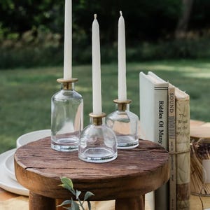 Puede incluir: Tres candelabros de vidrio transparente con bordes dorados, cada uno con una vela blanca alta. Los candelabros est&aacute;n colocados sobre una peque&ntilde;a mesa redonda de madera. Los libros est&aacute;n apilados a la derecha. El fondo es una escena borrosa al aire libre.