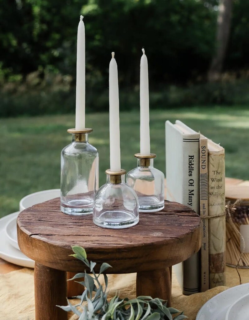 Puede incluir: Tres candelabros de vidrio transparente con bordes dorados, cada uno con una vela blanca alta. Los candelabros est&aacute;n colocados sobre una peque&ntilde;a mesa redonda de madera. Los libros est&aacute;n apilados a la derecha. El fondo es una escena borrosa al aire libre.