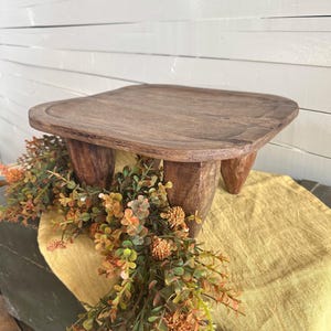May include: A dark brown wooden riser with a square top and four thick, tapered legs. The surface of the wood shows a natural grain pattern. The riser is positioned over a yellow fabric runner and a decorative garland of green and orange foliage.
