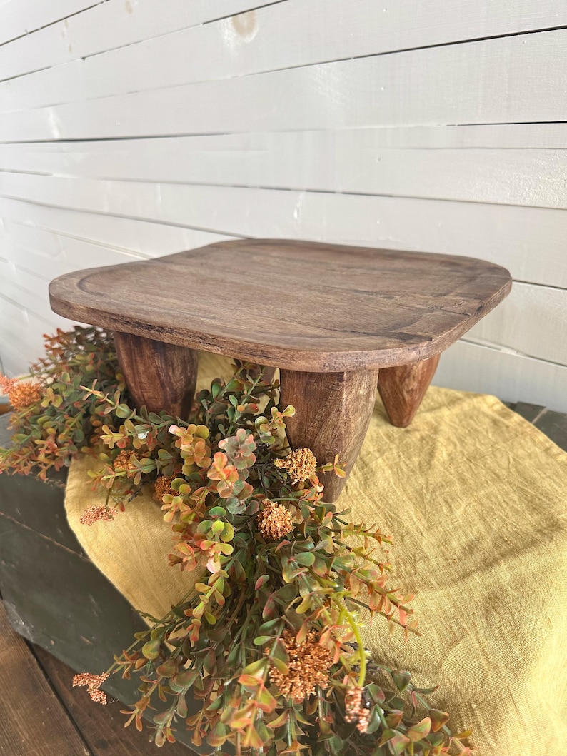 May include: A dark brown wooden riser with a square top and four thick, tapered legs. The surface of the wood shows a natural grain pattern. The riser is positioned over a yellow fabric runner and a decorative garland of green and orange foliage.