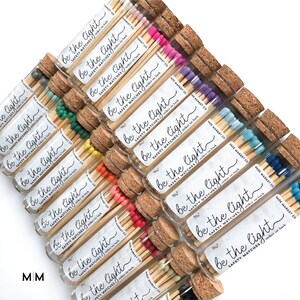 May include: A collection of glass tubes filled with safety matches, each capped with a cork. The match heads are various colors, including pink, purple, blue, and orange. Each tube has a label that reads "be the light".