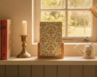 May include: A still-life scene featuring a wooden framed floral print, a brass candlestick with a white candle, and a small watering can with a floral design. The items are arranged on a white shelf near a window.