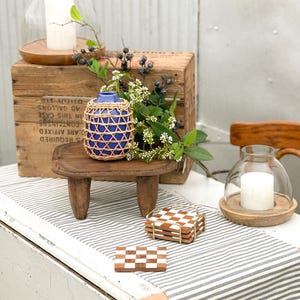 May include: A blue ceramic vase wrapped in a woven rattan cage, a small wooden stool with three legs, two glass candle domes with white pillar candles, and a stack of square brown and white checkered coasters in a gold metal holder.