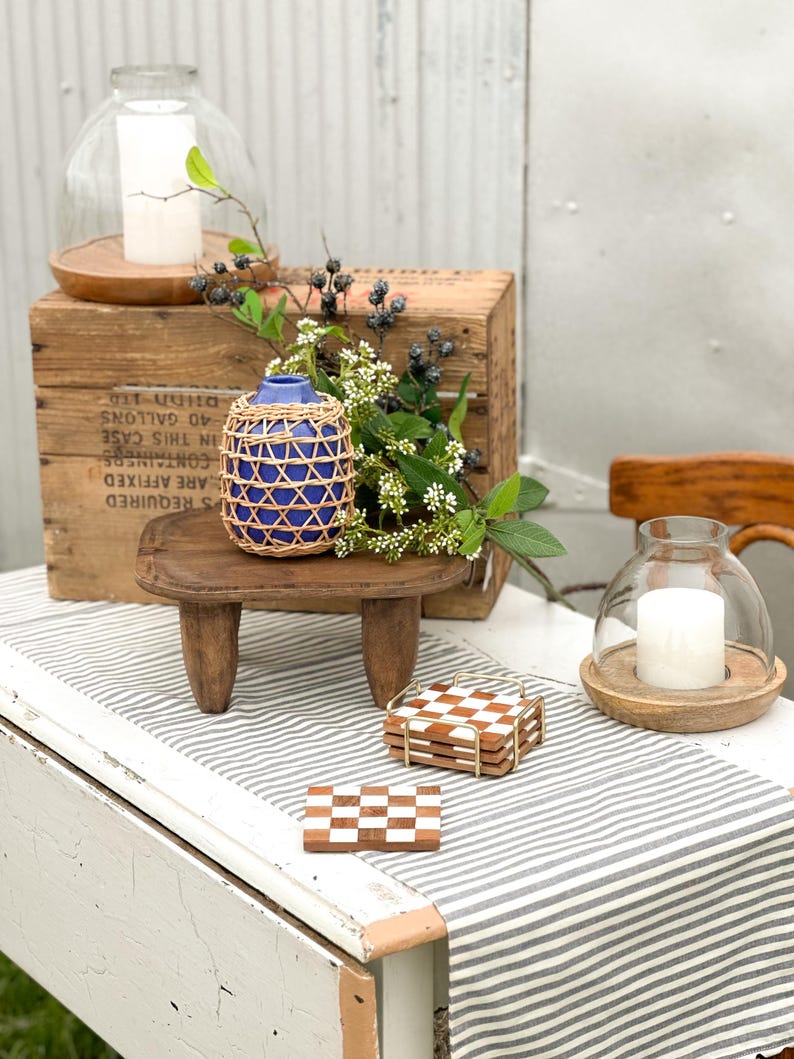 May include: A blue ceramic vase wrapped in a woven rattan cage, a small wooden stool with three legs, two glass candle domes with white pillar candles, and a stack of square brown and white checkered coasters in a gold metal holder.