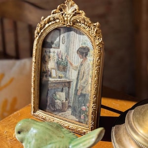 May include: Ornate gold-colored picture frame with a detailed, arched top. The frame holds a painting of a woman in a room. A green ceramic bird figurine sits in front of the frame on a wooden surface.