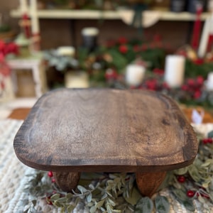May include: A square wooden riser with rounded corners and a dark brown finish, supported by four short, sturdy legs. The surface features a visible wood grain texture, providing a stable platform for display purposes.