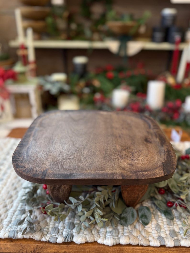 May include: A square wooden riser with rounded corners and a dark brown finish, supported by four short, sturdy legs. The surface features a visible wood grain texture, providing a stable platform for display purposes.
