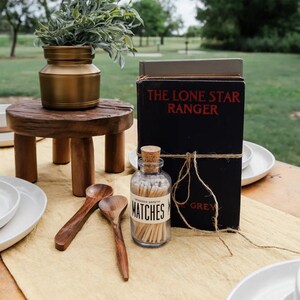 Puede incluir: Una mesa r&uacute;stica al aire libre con un peque&ntilde;o taburete de madera con un jarr&oacute;n dorado con vegetaci&oacute;n. Un libro negro titulado "The Lone Star Ranger" est&aacute; atado con hilo. Un tarro de cristal de cerillas y cucharas de madera completan la escena.