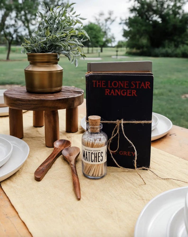Puede incluir: Una mesa r&uacute;stica al aire libre con un peque&ntilde;o taburete de madera con un jarr&oacute;n dorado con vegetaci&oacute;n. Un libro negro titulado "The Lone Star Ranger" est&aacute; atado con hilo. Un tarro de cristal de cerillas y cucharas de madera completan la escena.